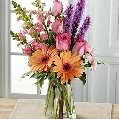 Exuding a special charm, with a casual fresh-from-the-garden look, this gorgeous spring bouquet is the perfect way to delight your recipient in honor of any of life's most treasured moments. Peach gerbera daisies are soft and sophisticated surrounded by pink roses, pink snapdragons, pink mini carnations, purple liatris, and lush greens arranged with an artist's eye in a gathered square clear glass vase.